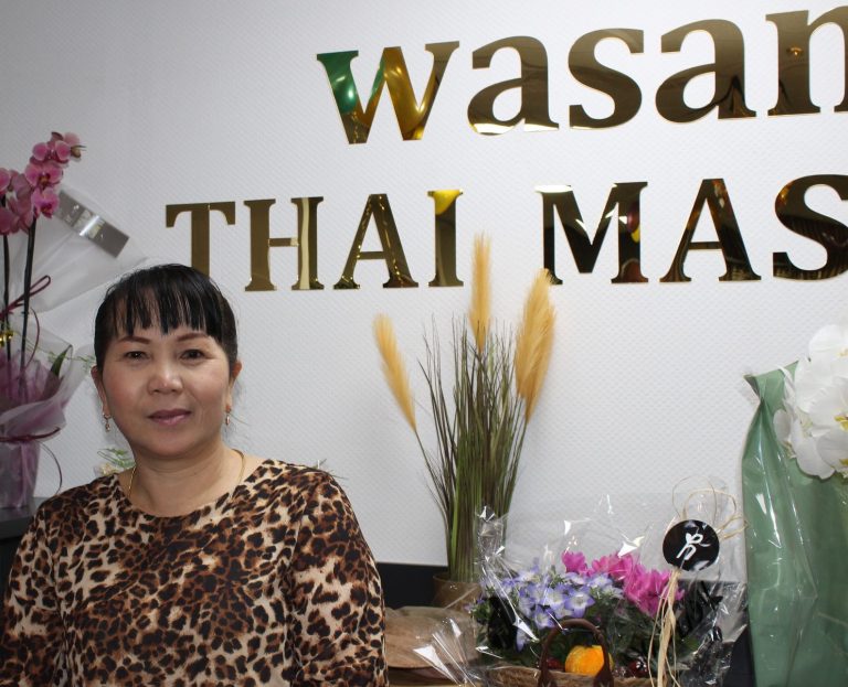 Bild von Wasana Wasana mit buntem Oberteil steht vor einer Wand mit dem Schriftzug "Wasan Thai Massage".